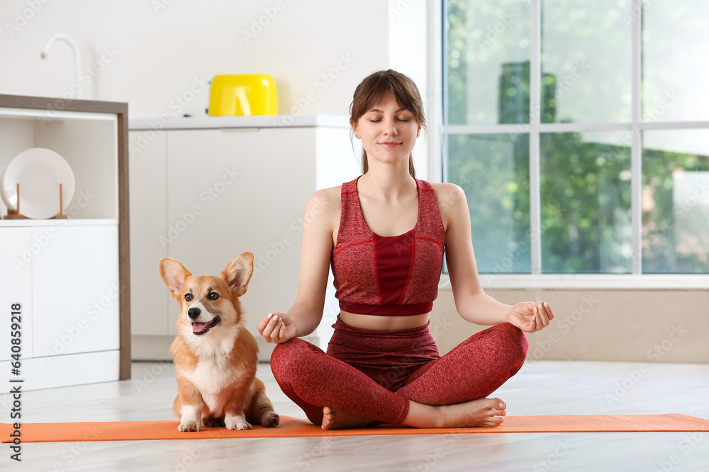 Sporty young woman meditating with cute Corgi dog on yoga mat in kitchen