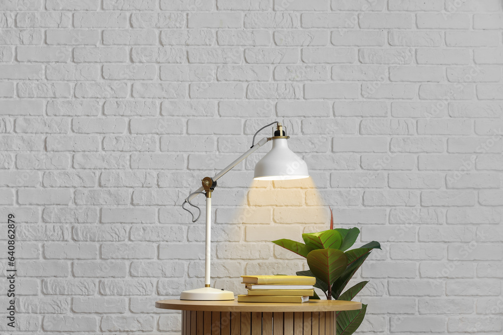 Table with glowing lamp, books and houseplant near white brick wall in room