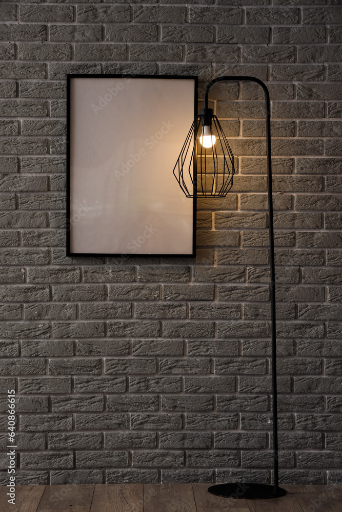 Glowing lamp and blank frame near grey brick wall