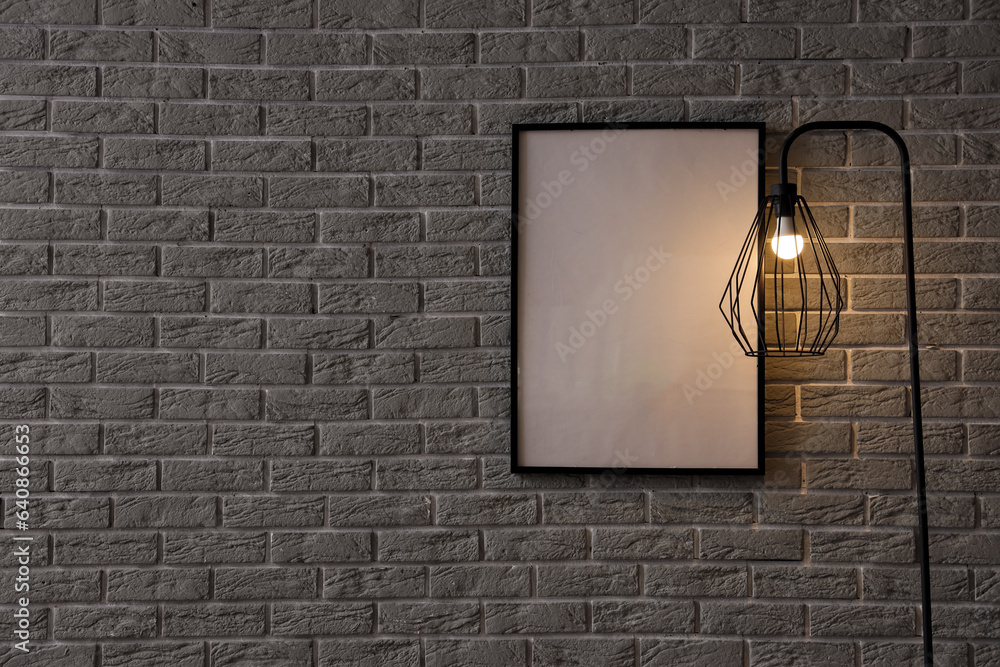 Glowing lamp and blank frame near grey brick wall