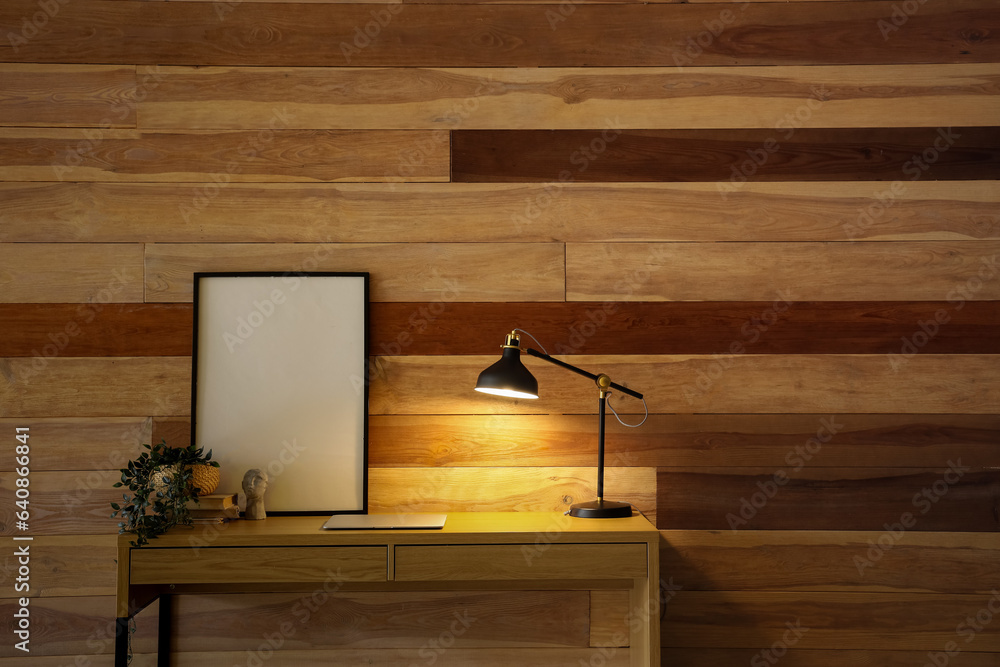 Desk with glowing lamp, modern laptop, blank frame and houseplant near wooden wall