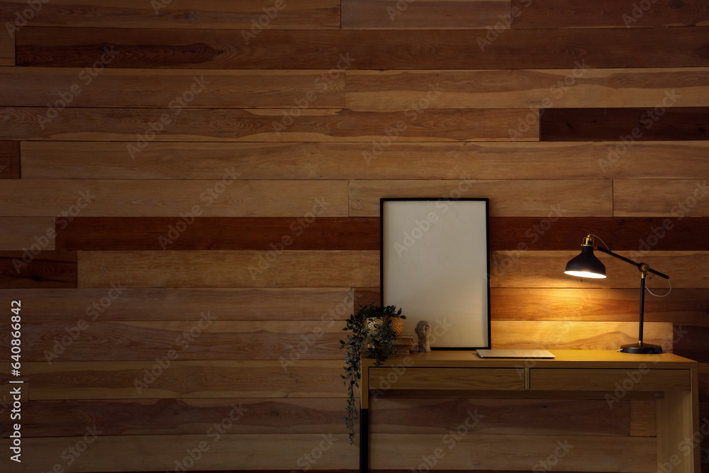 Desk with glowing lamp, modern laptop, blank frame and houseplant near wooden wall