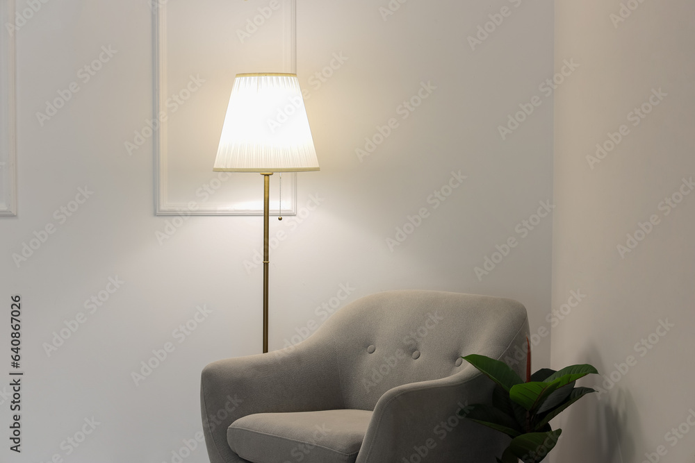 Glowing lamp, grey armchair and houseplant near white wall