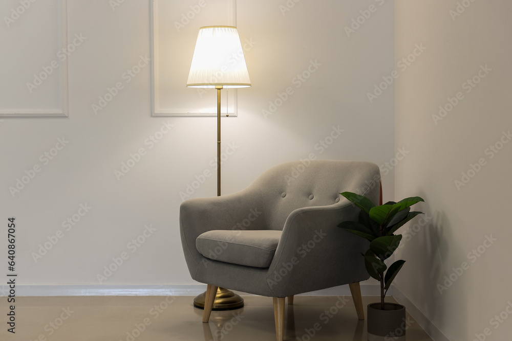 Glowing lamp, grey armchair and houseplant near white wall