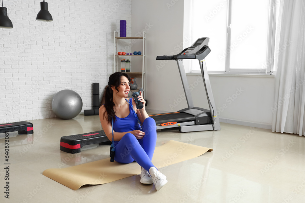 Sporty young woman with jumping rope drinking water in gym