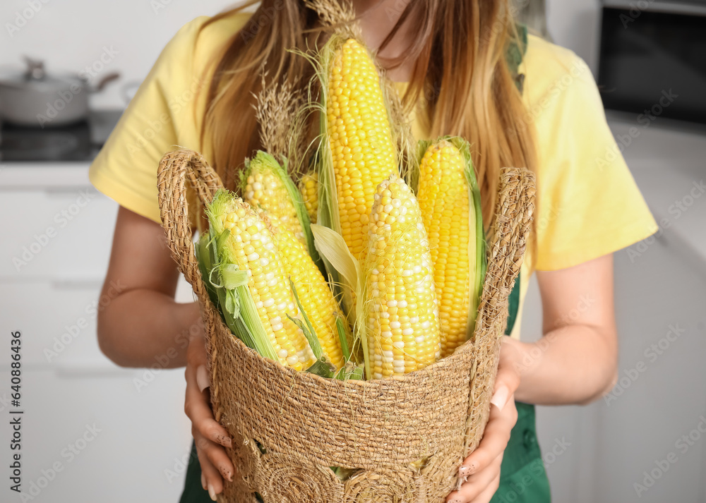 Beautiful young female farmer with wicker basket full of ripe corn cobs in kitchen