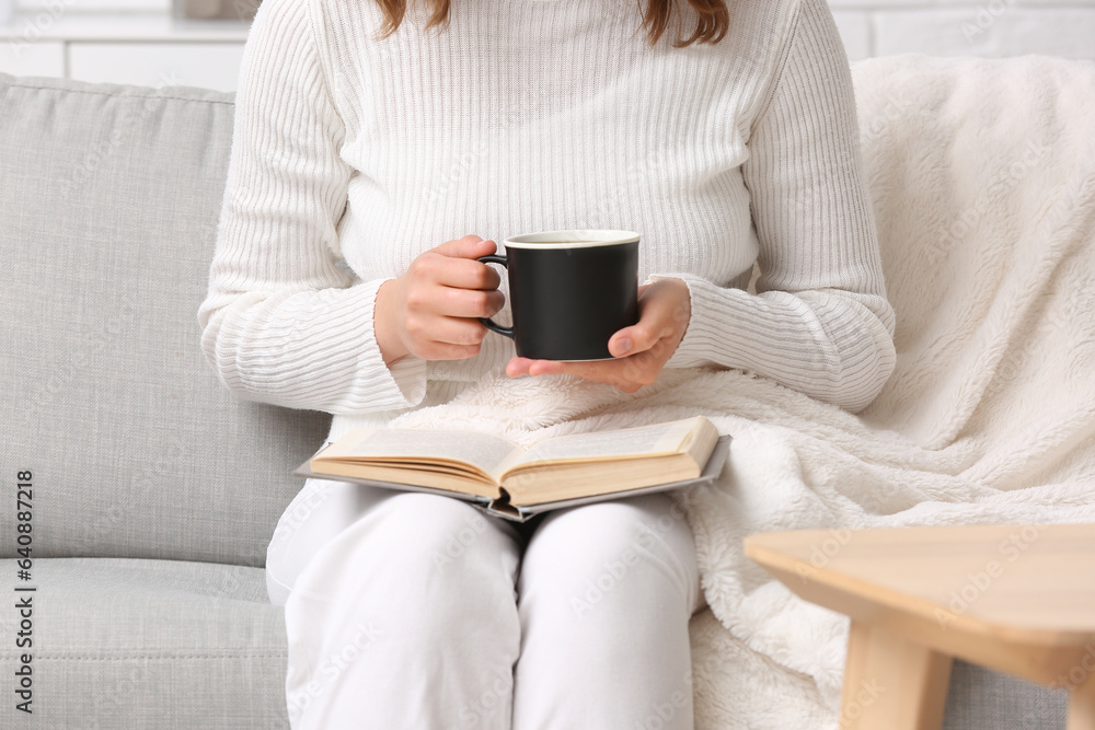 Woman with cup of tasty coffee and book resting on sofa at home