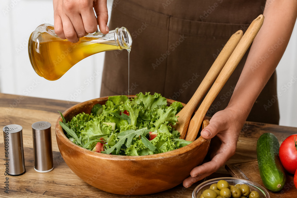 Woman adding olive oil into bowl with tasty salad at table in kitchen