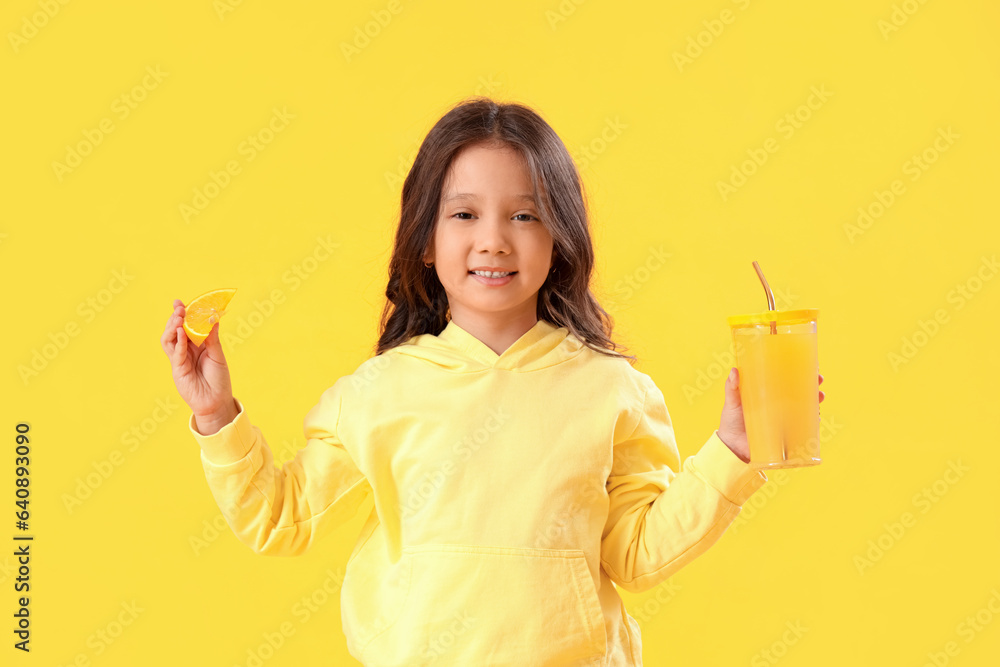 Little Asian girl with cup of fresh citrus juice and orange slice on yellow background