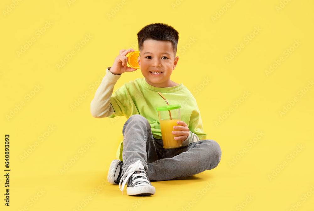 Little Asian boy with cup of fresh citrus juice and orange slice sitting on yellow background