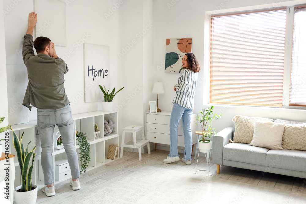 Young couple hanging paintings on light wall at home