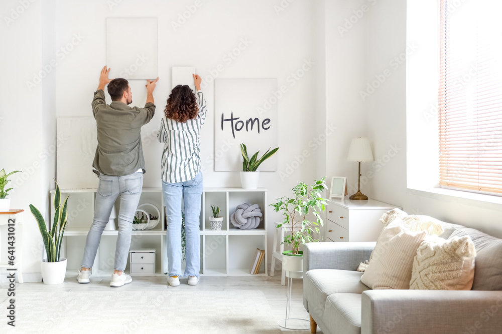 Young couple hanging paintings on light wall at home, back view