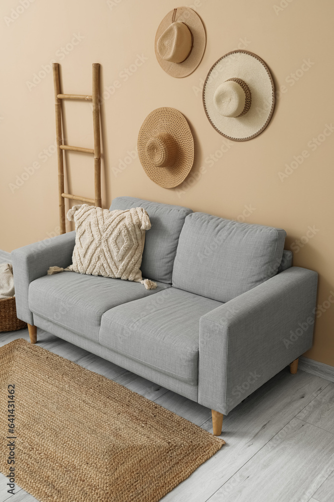 Cozy grey sofa with cushion, ladder and hanging hats near beige wall