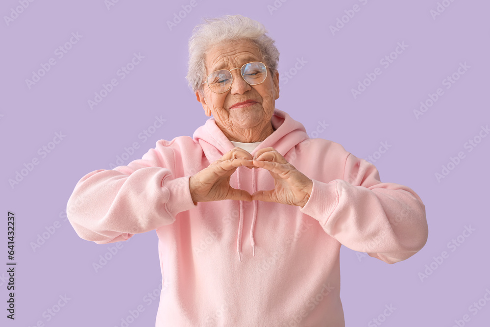 Senior woman making heart with her hands on lilac background