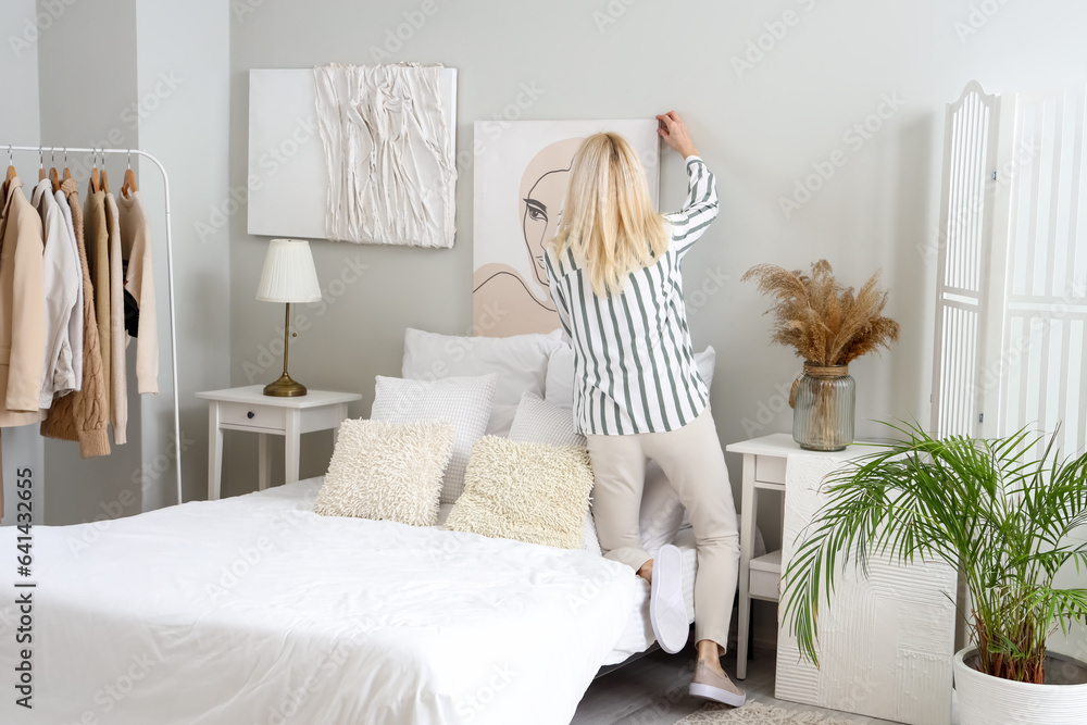 Mature woman hanging painting on light wall in bedroom, back view