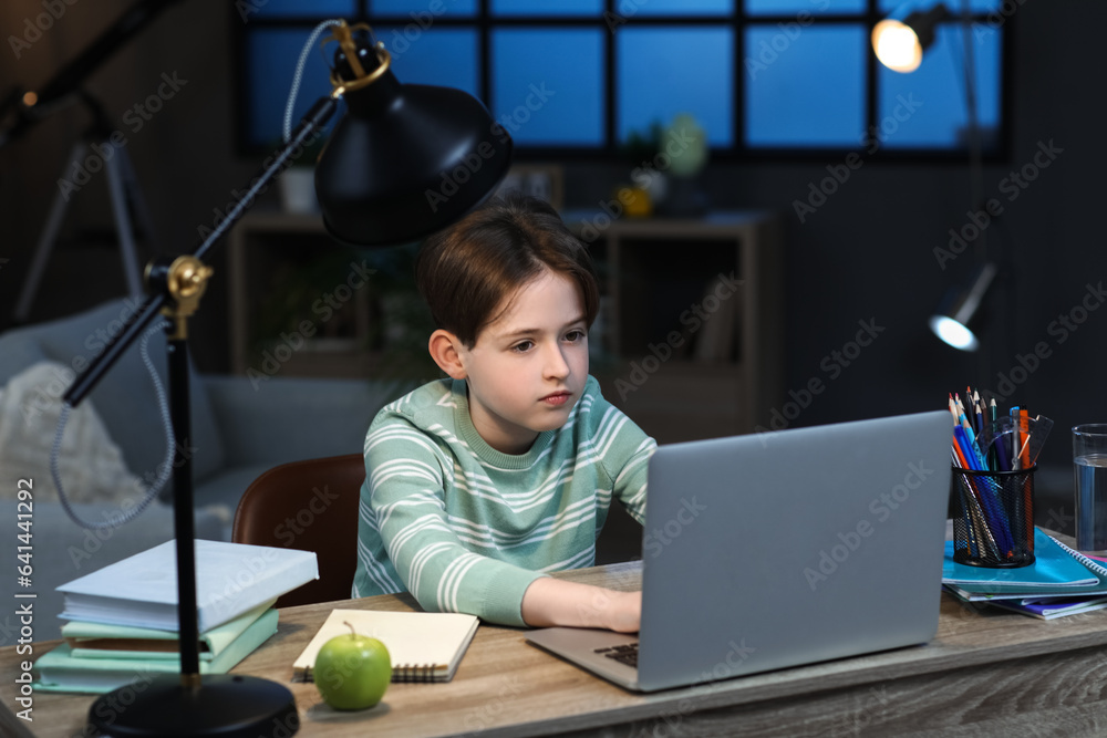 Little boy with laptop doing homework at home late in evening