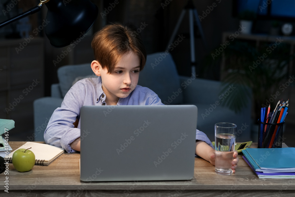 Little boy doing homework with laptop and taking glass of water at home late in evening