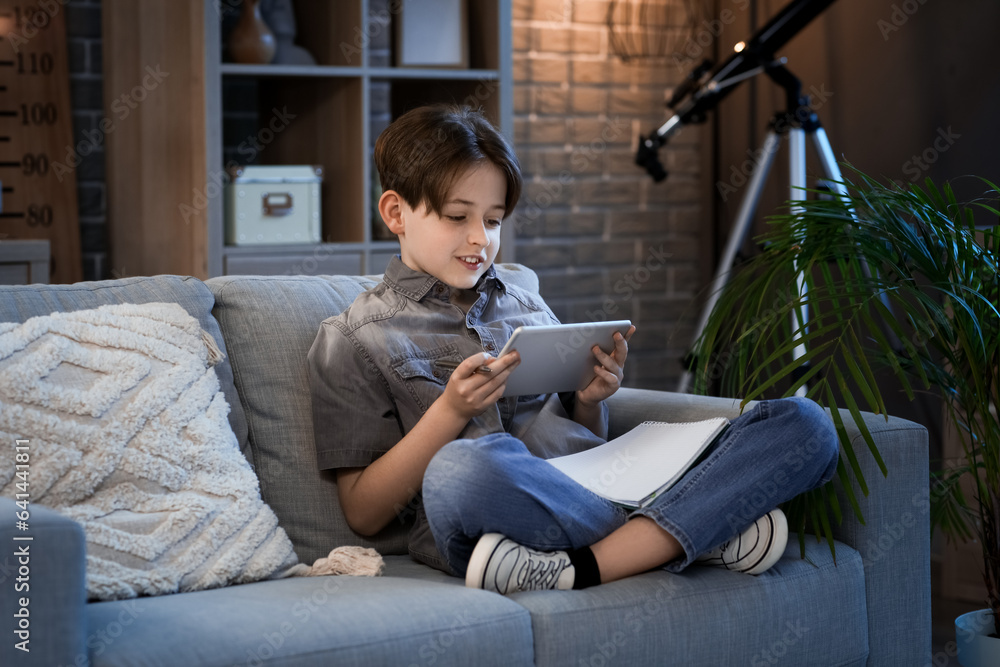 Little boy doing homework with tablet and sitting on sofa at home in evening