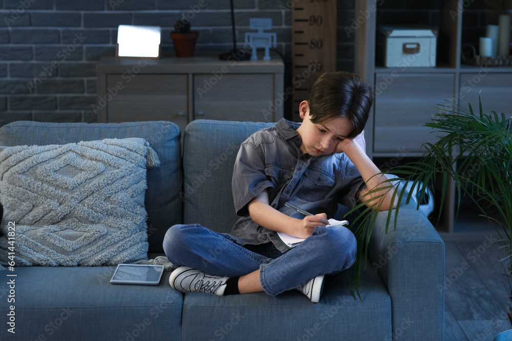 Little boy doing homework with tablet and sitting on sofa at home in evening