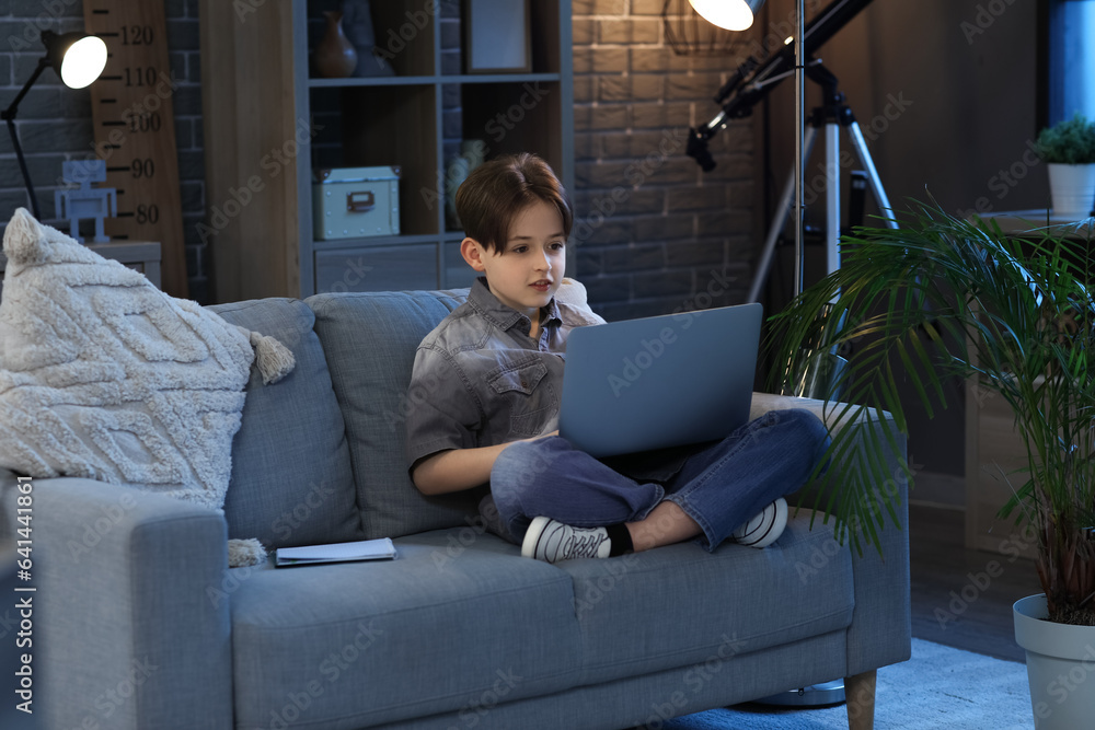 Little boy doing homework with laptop and sitting on sofa at home in evening