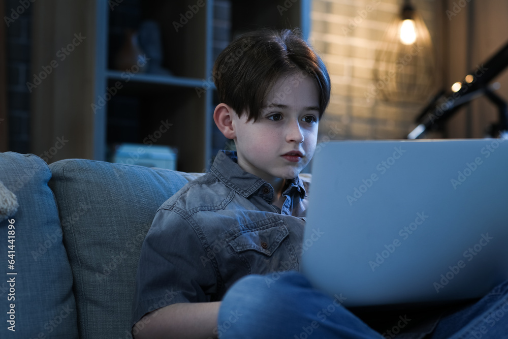 Little boy doing homework with laptop and sitting on sofa at home in evening