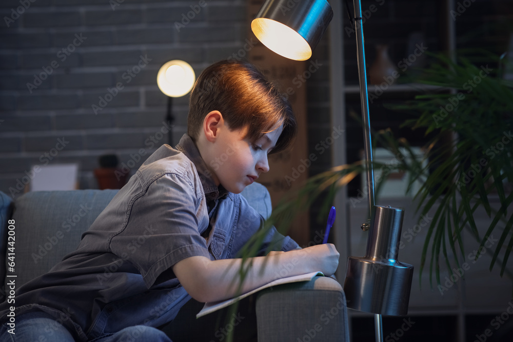 Little boy sitting on sofa and doing homework at home in evening