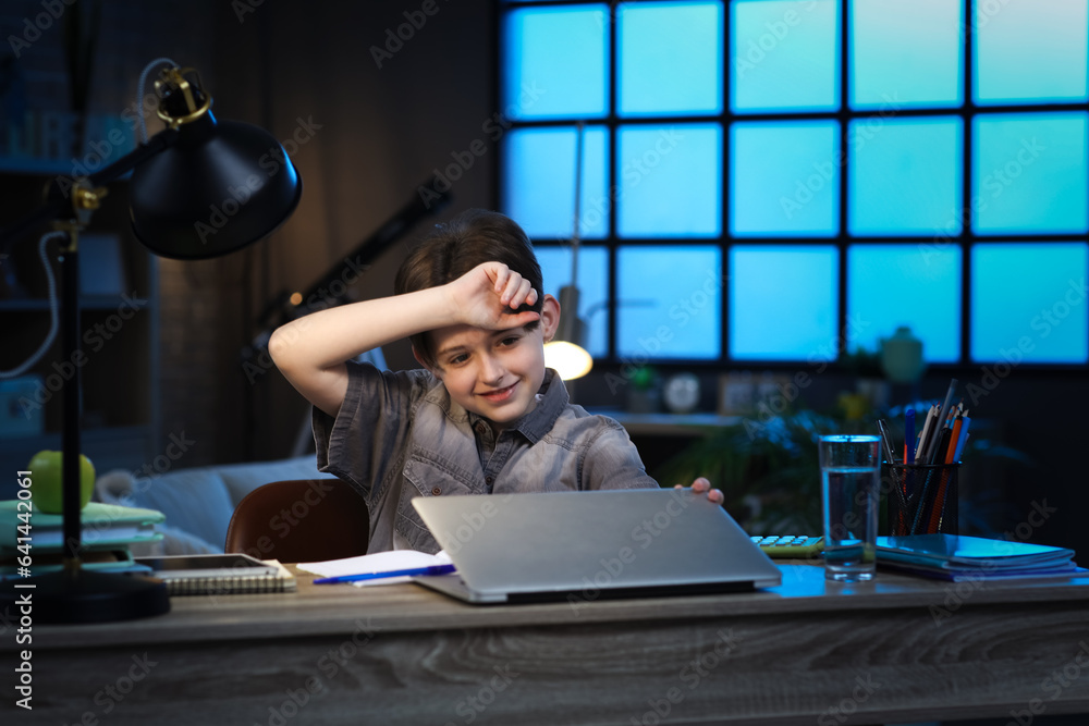 Tired little boy doing homework with laptop at home late in evening