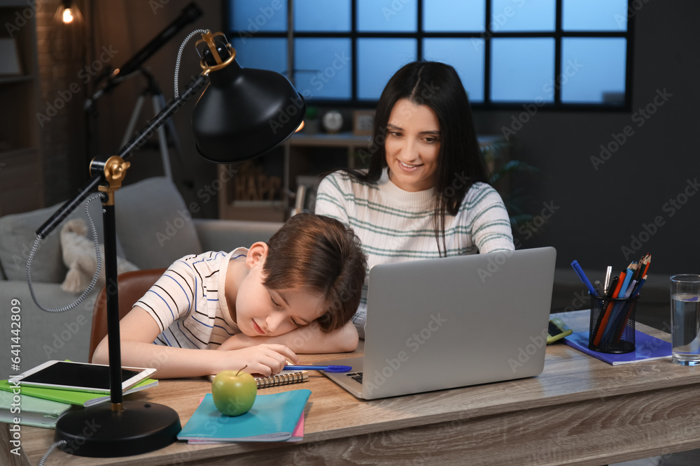 Mother with her little sleeping son at table doing homework late in evening