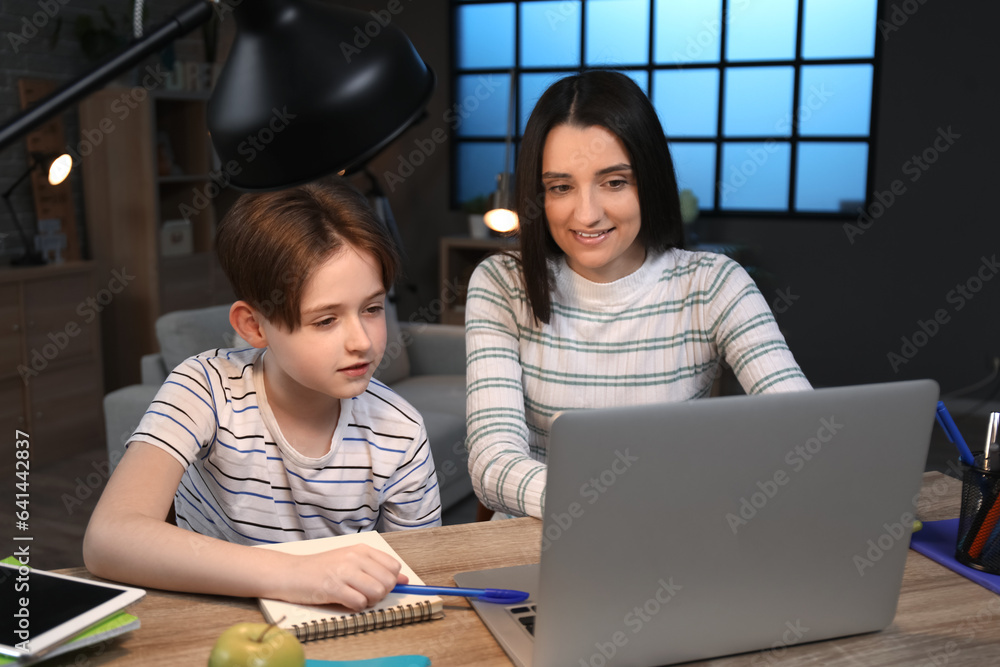 Mother with her little son sitting at table and doing homework late in evening