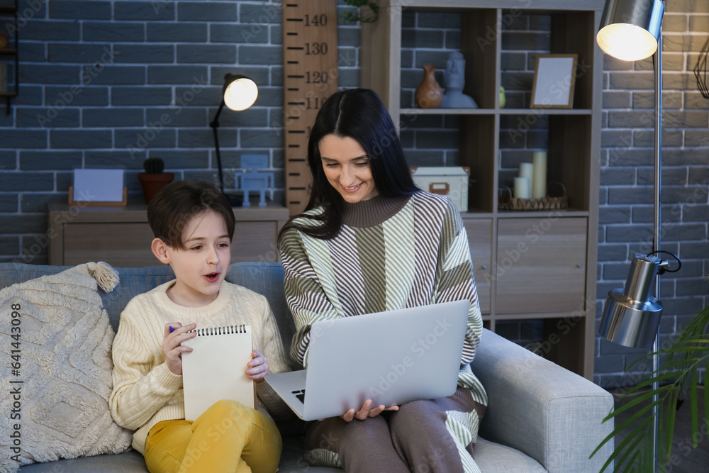 Little boy and his mother doing homework with laptop at home late in evening