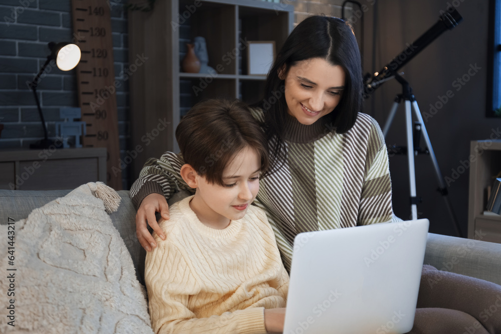 Little boy and his mother doing homework with laptop at home late in evening