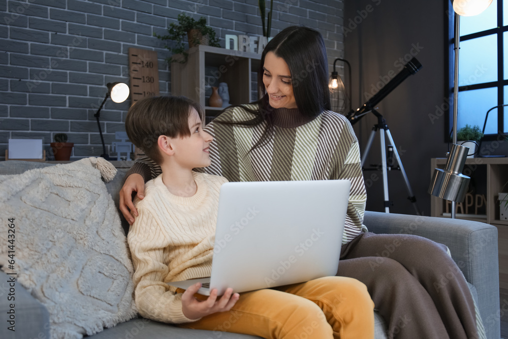 Little boy and his mother doing homework with laptop at home late in evening