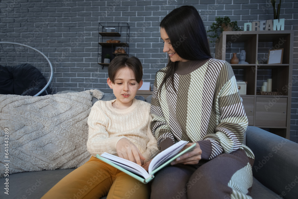 Little boy with his mother sitting on sofa and reading book at home late in evening