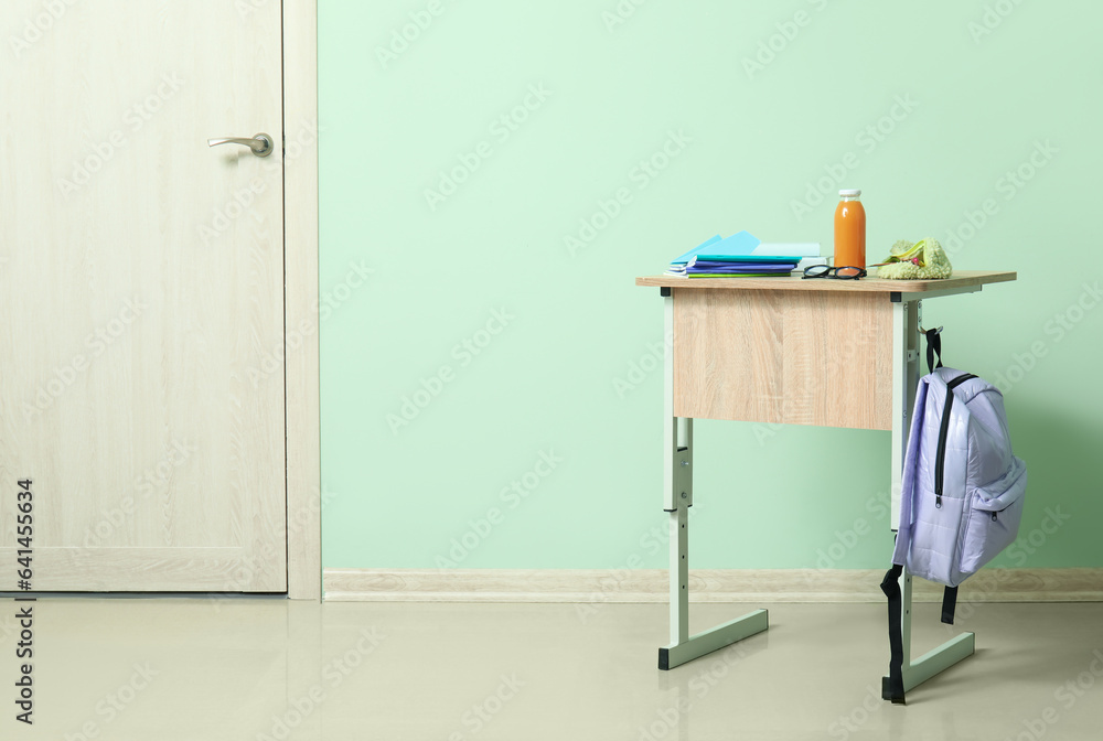 Modern school desk with backpack, bottle of juice and stationery in room near green wall