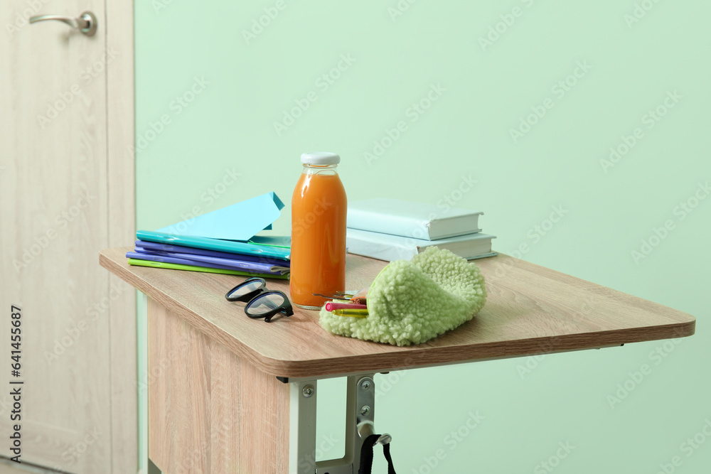 Modern school desk with backpack, bottle of juice and stationery in room near green wall