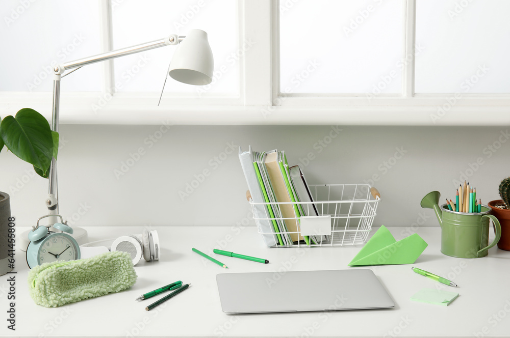 Modern school desk with laptop and stationery in room near white wall