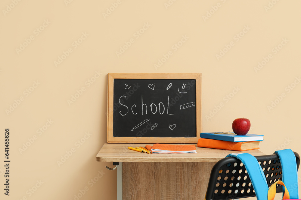 Modern school desk, backpack, stationery and blackboard with word SCHOOL in room near beige wall