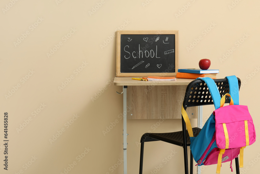 Modern school desk, backpack, stationery and blackboard with word SCHOOL in room near beige wall