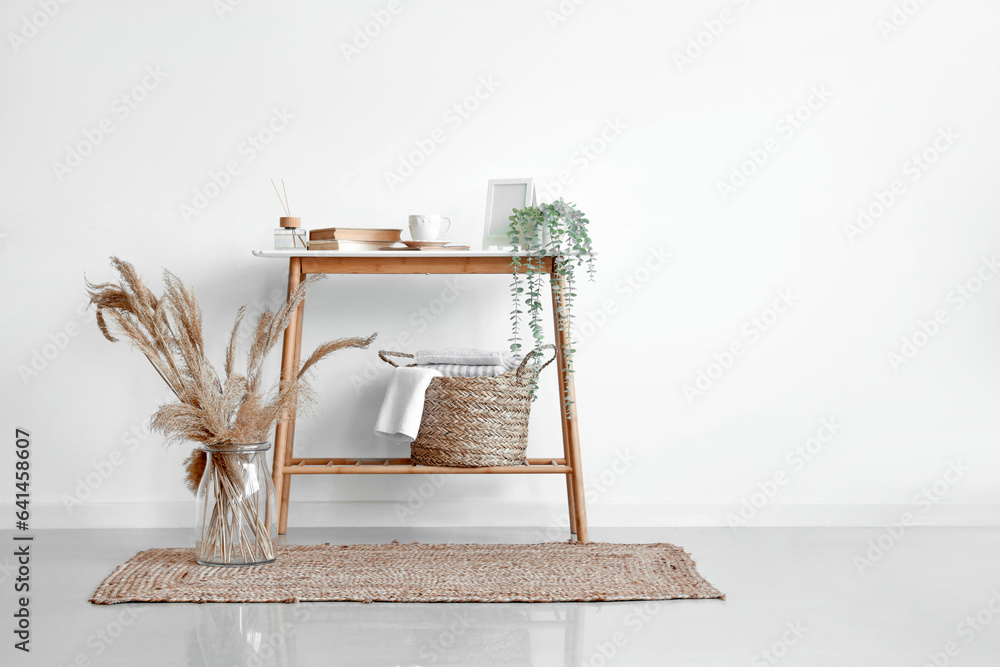 Wicker basket with towels on console table and pampas grass in vase near white wall