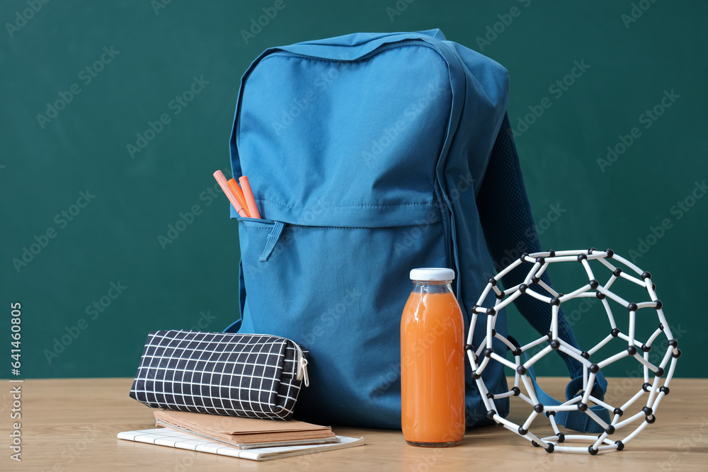 Blue school backpack with stationery and bottle of juice on wooden table near green chalkboard