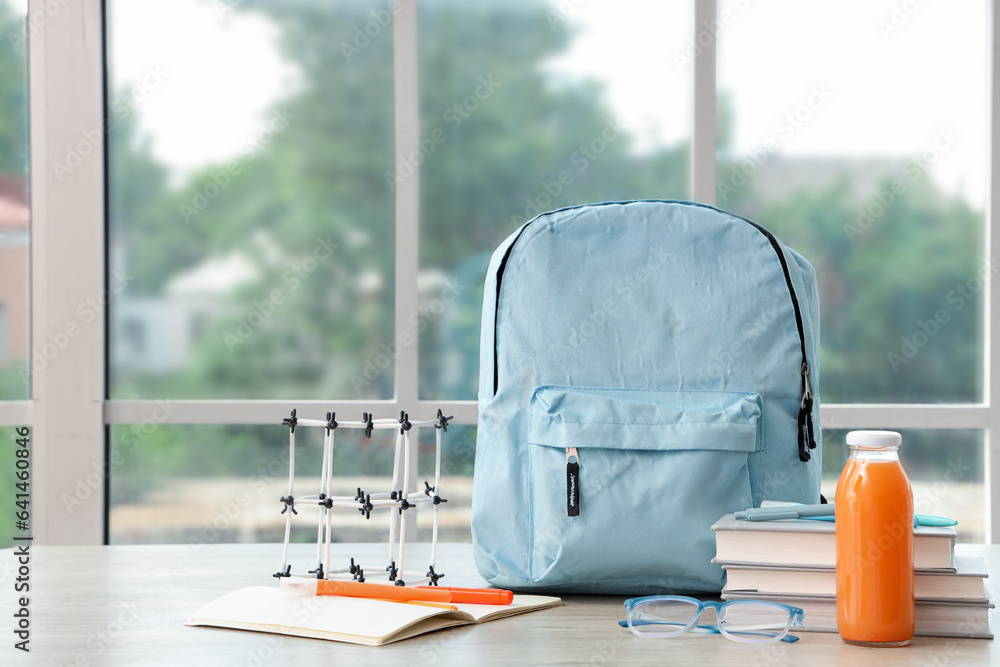 Blue school backpack with stationery, molecular model and bottle of juice on wooden table near windo