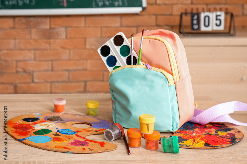 Stylish school backpack with art supplies on desk in classroom