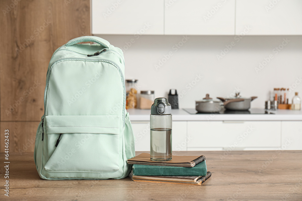 Stylish school backpack with stationery and bottle of water on table in kitchen