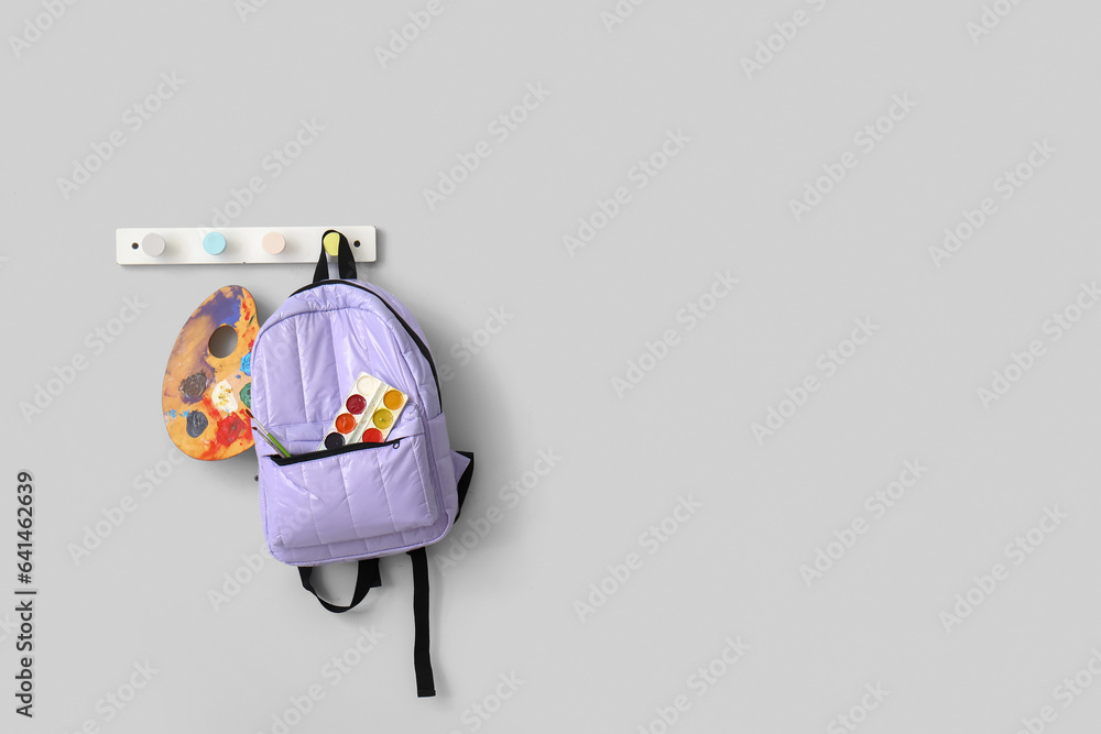 School backpack with paints and brushes hanging on light wall