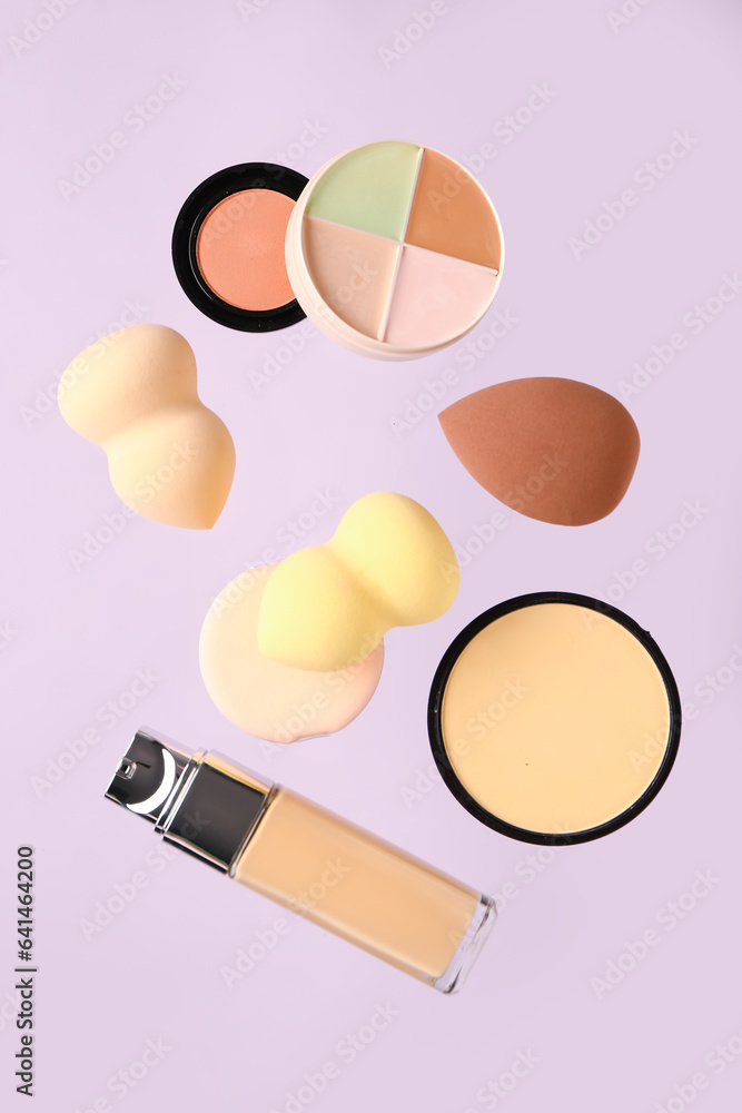 Makeup sponges and decorative cosmetics flying on lilac background