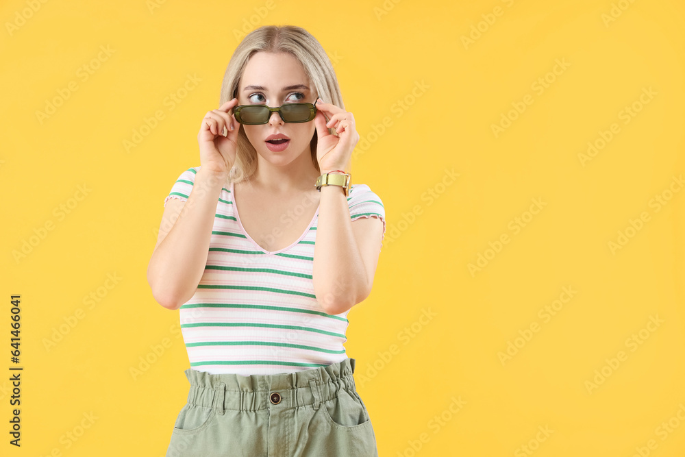 Shocked young beautiful woman in stylish sunglasses on yellow background