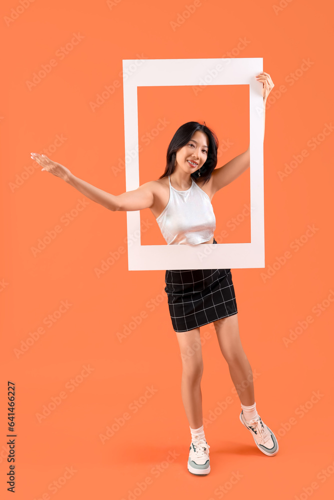 Young beautiful Asian woman with frame pointing at something on orange background