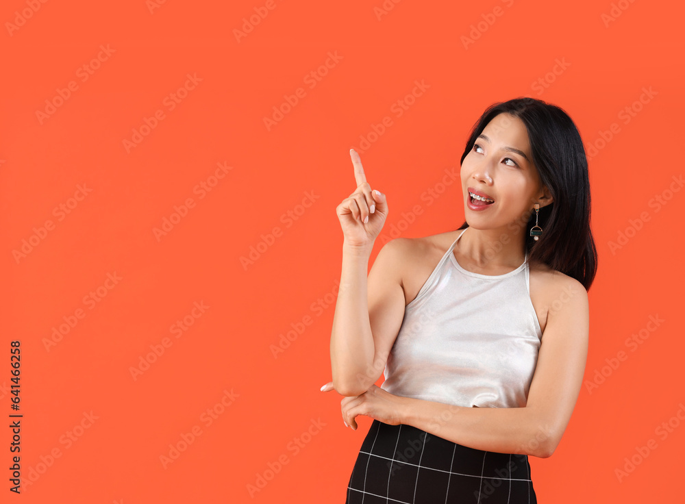 Happy young beautiful Asian woman pointing at something on orange background