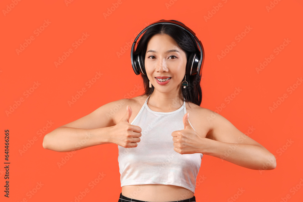 Happy young beautiful Asian woman in headphones showing thumbs-up gesture on orange background