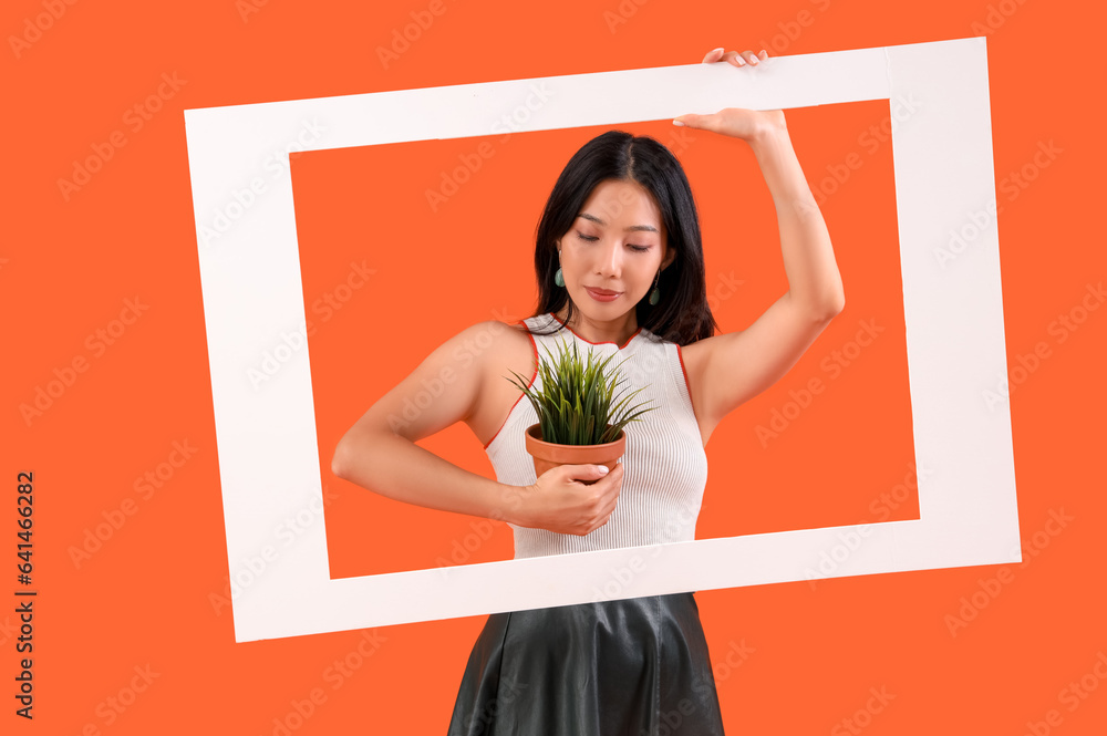 Young beautiful Asian woman with frame and houseplant on orange background